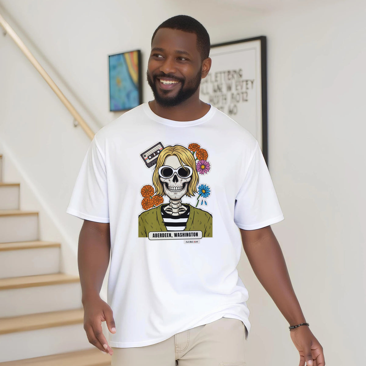 Uomo con la barba sorride vicino a una scala di legno, in casa, e indossa la t-shirt bianca con la calavera Aberdeen