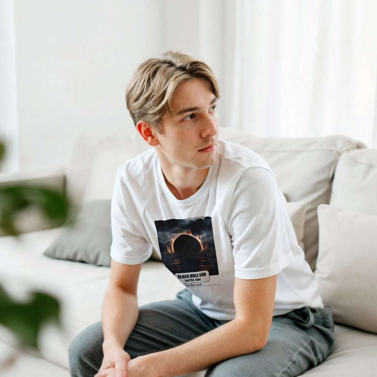 Ragazzo biondo seduto su un divano bianco, piegato in avanti, indossa la t-shirt bianca con la stampa Rock Art «Black Hole Sun – Seattle, 1994»