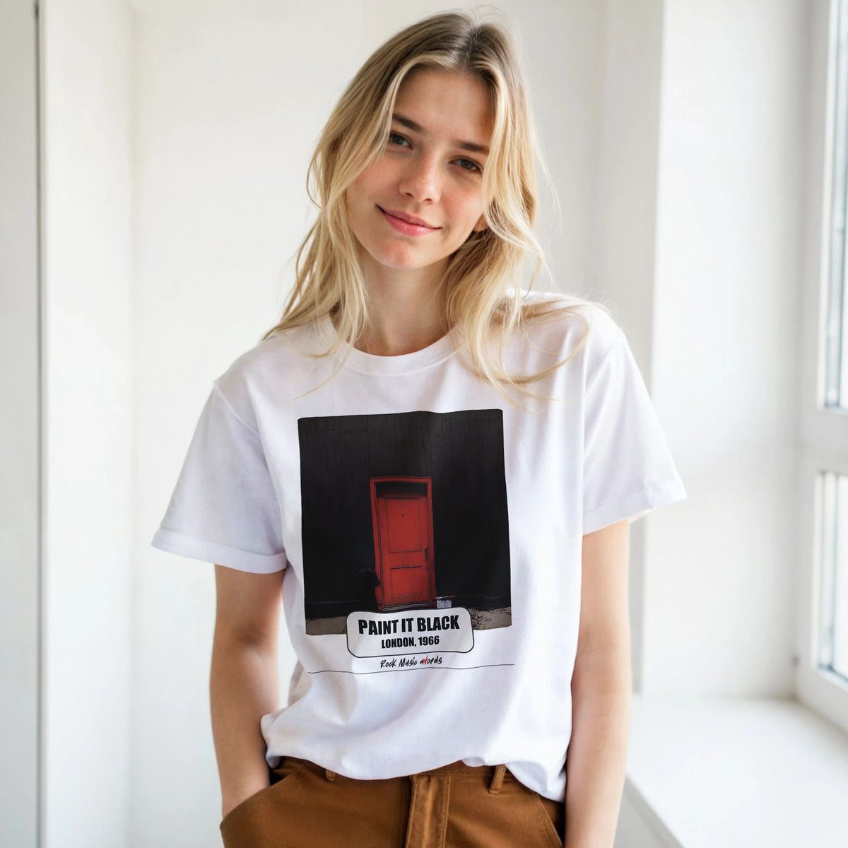 Ragazza bionda con le mani in tasca, accanto a una finestra, indossa la t-shirt bianca con la stampa Rock Art «Paint It Black – London, 1966»