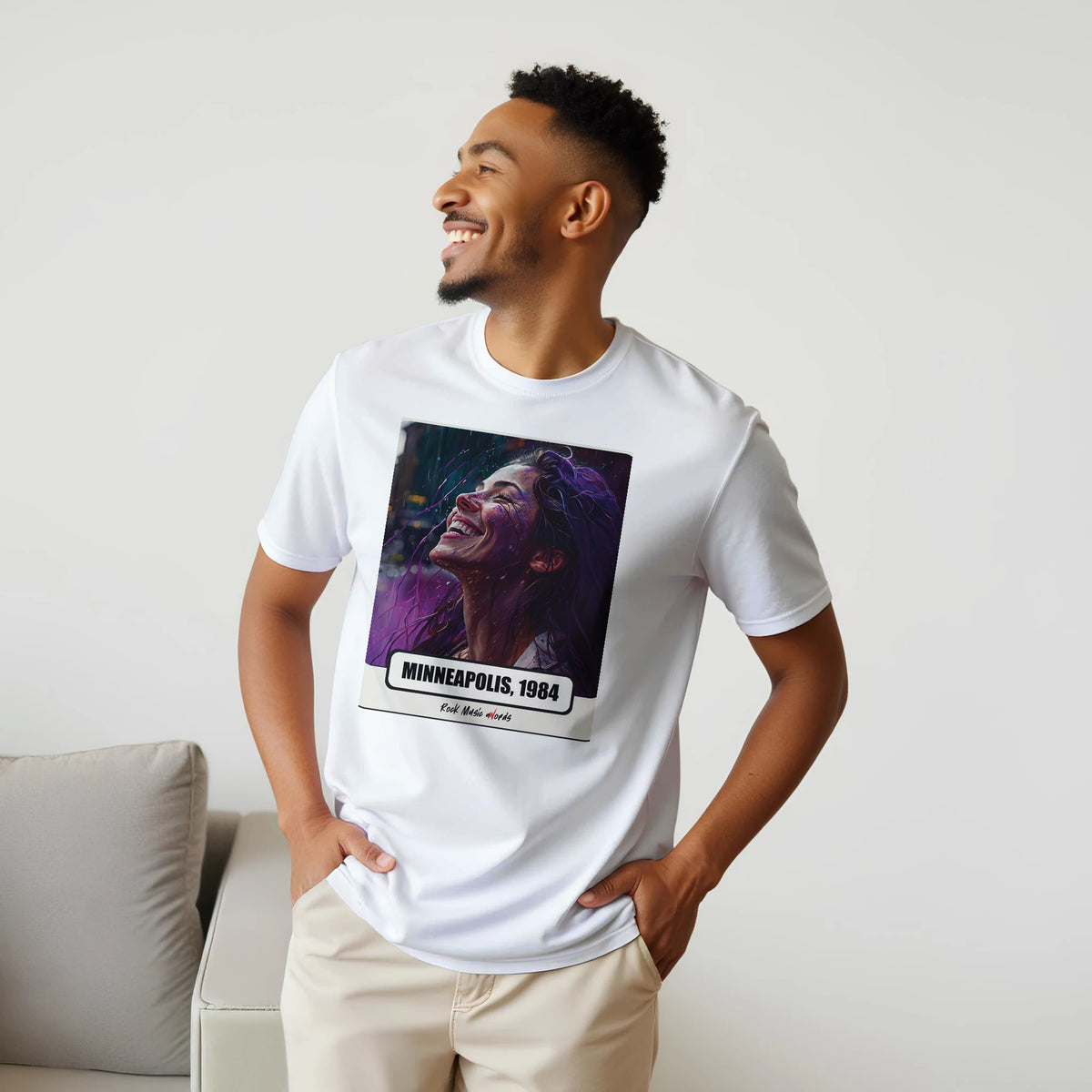 Uomo sorridente con le mani sui fianchi, accanto a un divano, indossa la t-shirt bianca con la stampa Rock Art «Purple Rain – Minneapolis, 1984»