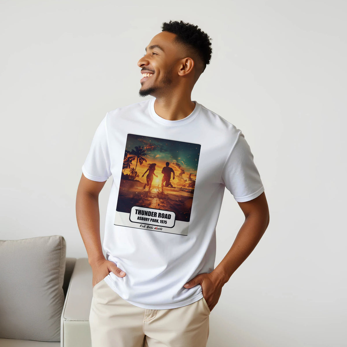 Uomo sorridente con le mani sui fianchi, accanto a un divano, indossa la t-shirt bianca con la stampa Rock Art «Thunder Road – Asbury Park, 1975»