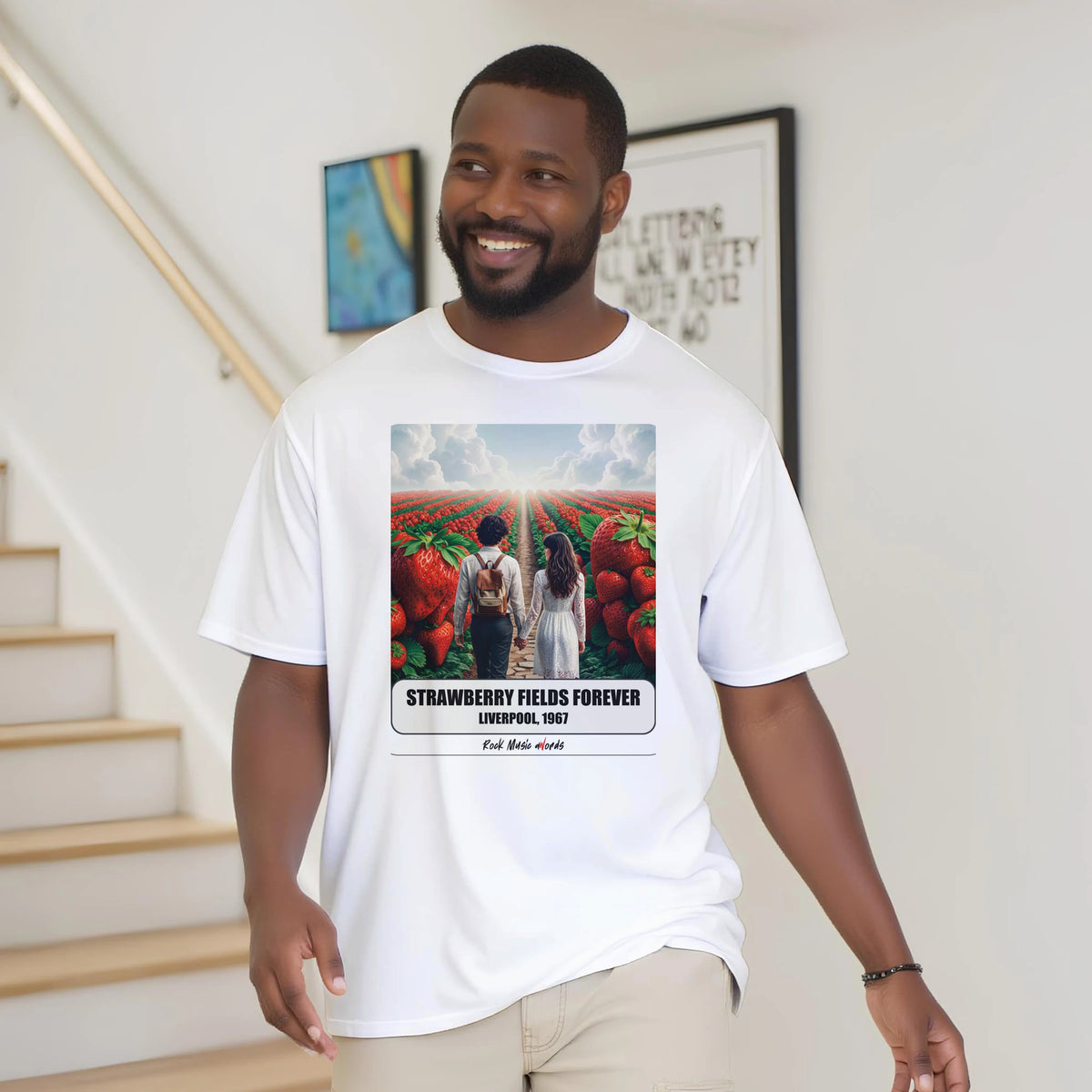 Uomo con la barba sorride vicino a una scala di legno, in casa, e indossa la t-shirt bianca con la stampa Rock Art «Strawberry Fields Forever – Liverpool, 1967»