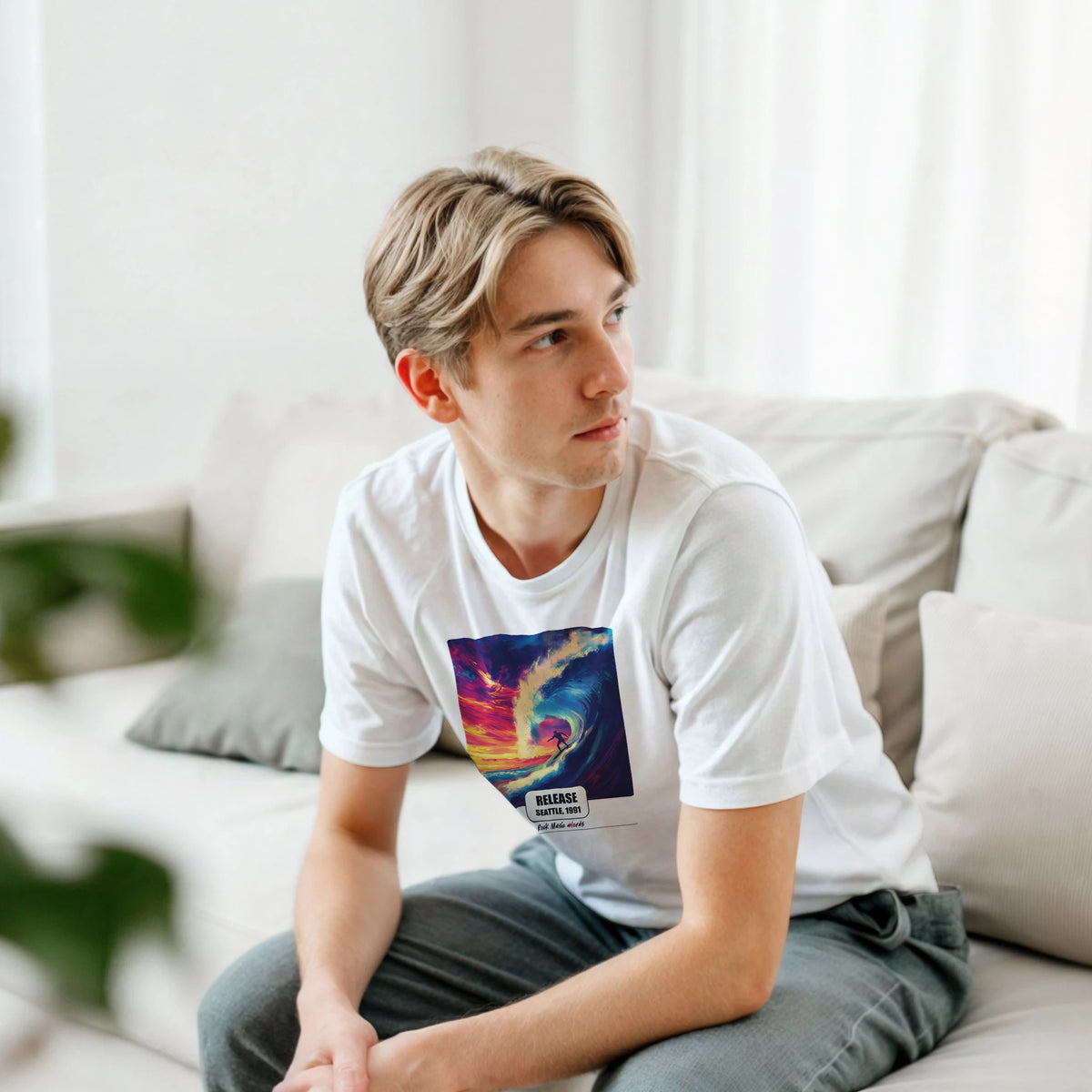 Ragazzo biondo seduto su un divano bianco, piegato in avanti, indossa la t-shirt bianca con la stampa Rock Art «Release – Seattle, 1991»