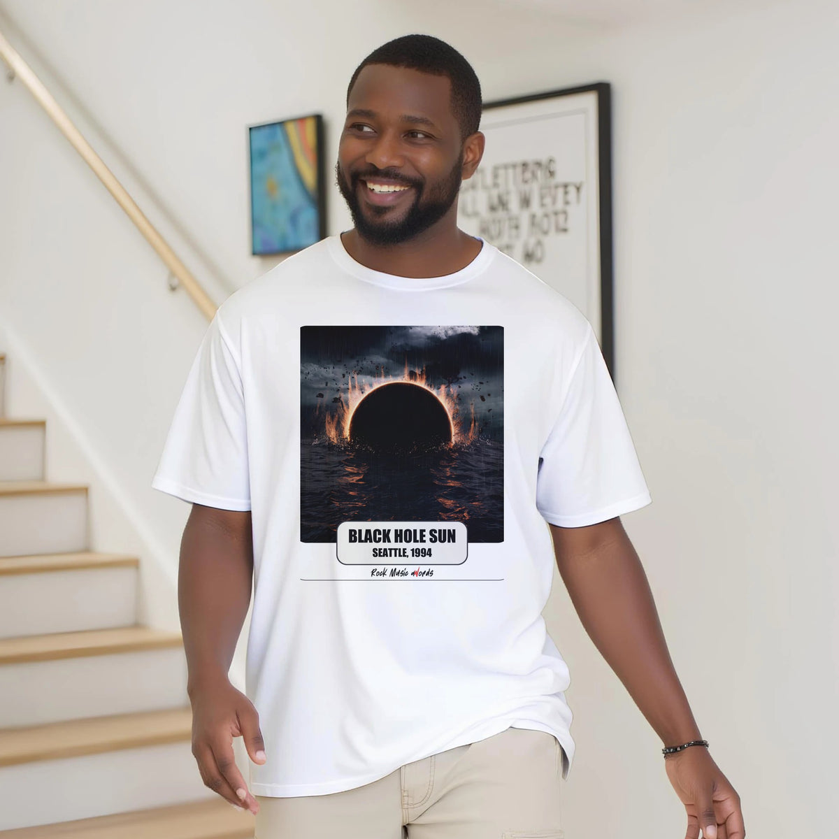 Uomo con la barba sorride vicino a una scala di legno, in casa, e indossa la t-shirt bianca con la stampa Rock Art «Black Hole Sun – Seattle, 1994»