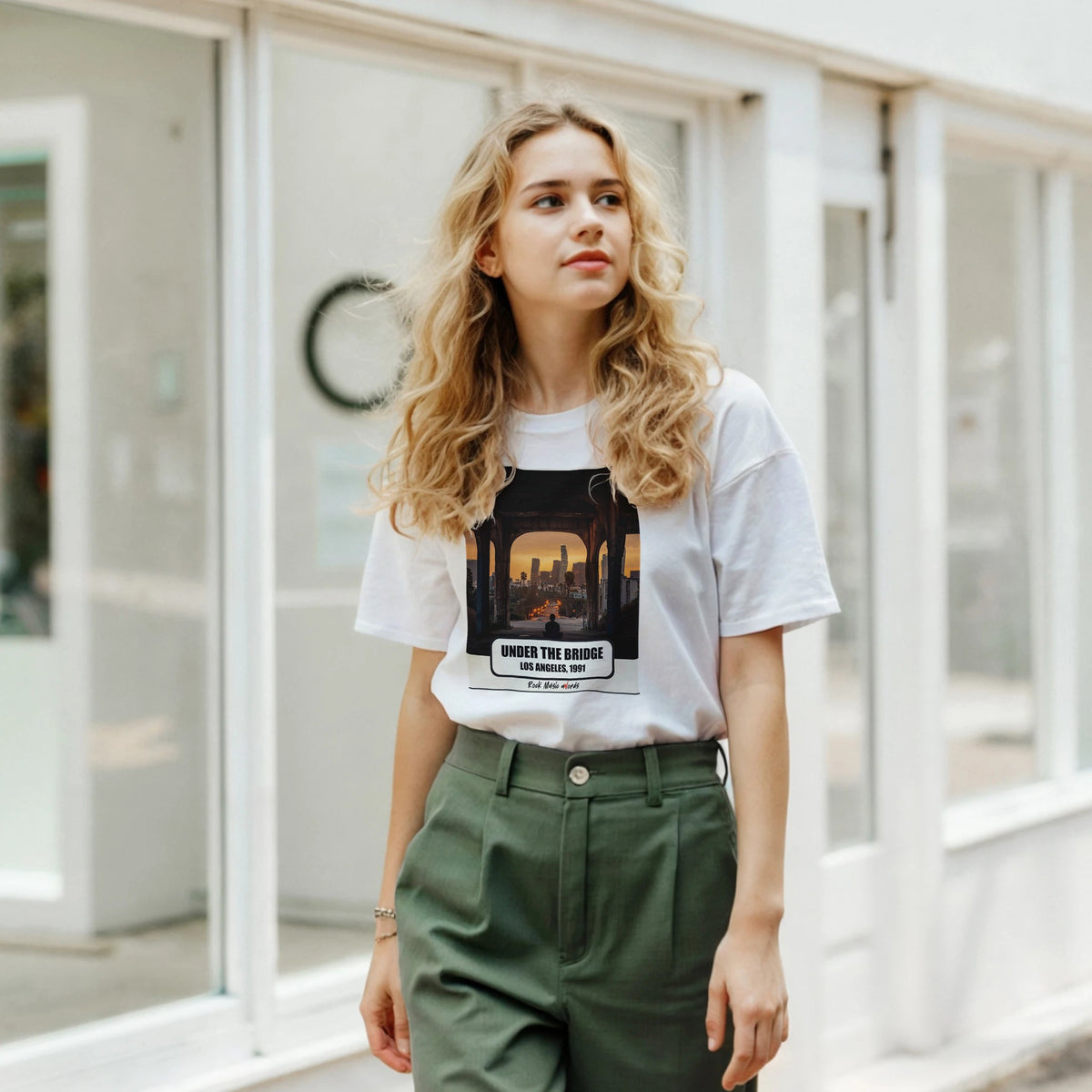 Ragazza bionda davanti a una facciata a vetrate indossa la t-shirt bianca con la stampa Rock Art «Under the Bridge – Los Angeles, 1991» e pantaloni verdi