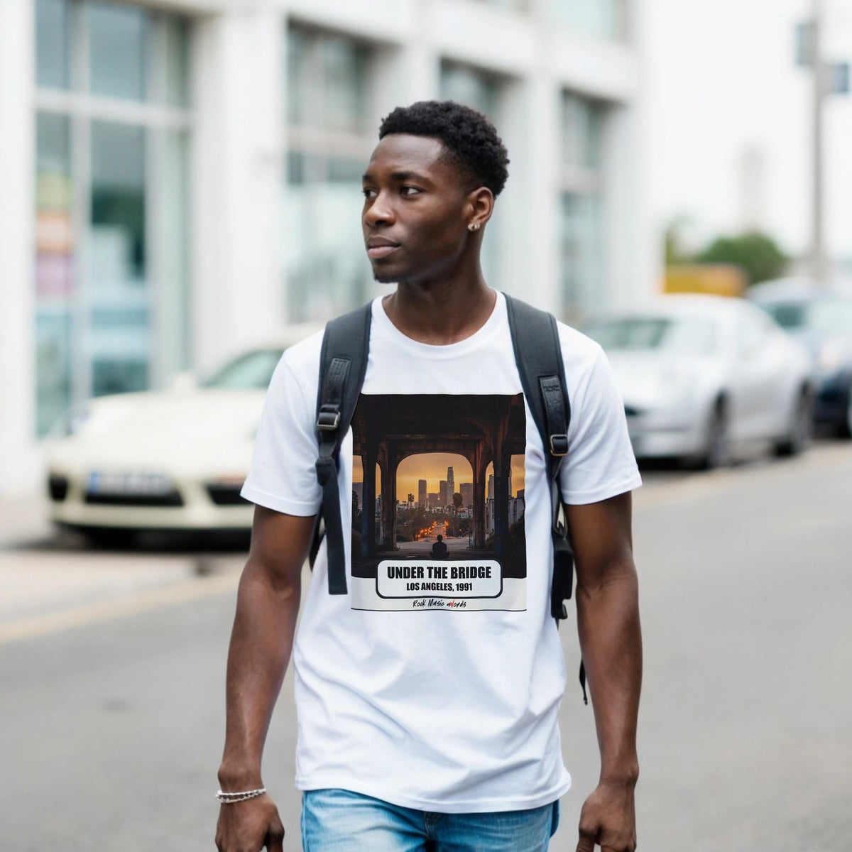 Ragazzo con zaino nero e jeans cammina per una via di città indossando la t-shirt bianca con la stampa Rock Art «Under the Bridge – Los Angeles, 1991»