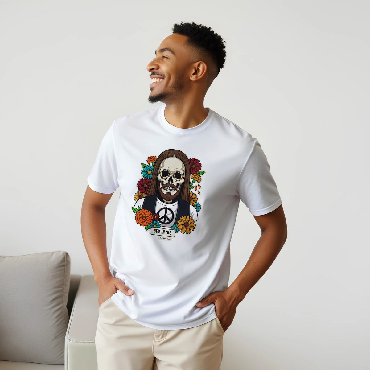 Uomo sorridente con le mani sui fianchi, accanto a un divano, indossa la t-shirt bianca con la calavera Bed-In '69