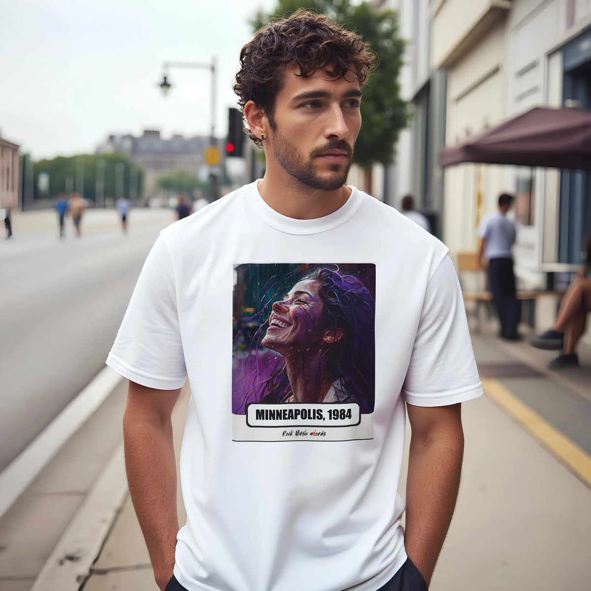 Uomo con capelli ricci e barba su un marciapiede di città indossa la t-shirt bianca con la stampa Rock Art «Purple Rain – Minneapolis, 1984»