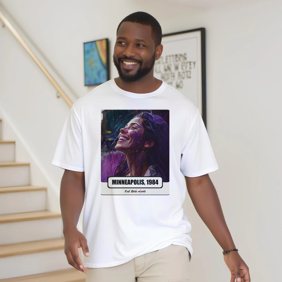Uomo con la barba sorride vicino a una scala di legno, in casa, e indossa la t-shirt bianca con la stampa Rock Art «Purple Rain – Minneapolis, 1984»