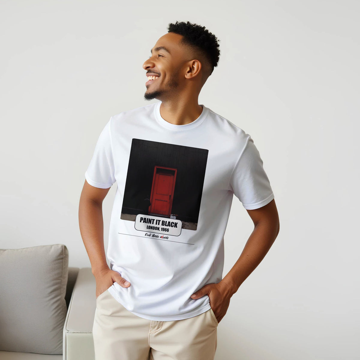 Uomo sorridente con le mani sui fianchi, accanto a un divano, indossa la t-shirt bianca con la stampa Rock Art «Paint It Black – London, 1966»