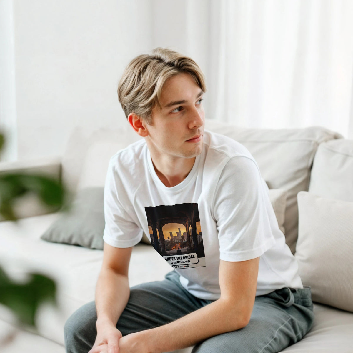 Ragazzo biondo seduto su un divano bianco, piegato in avanti, indossa la t-shirt bianca con la stampa Rock Art «Under the Bridge – Los Angeles, 1991»