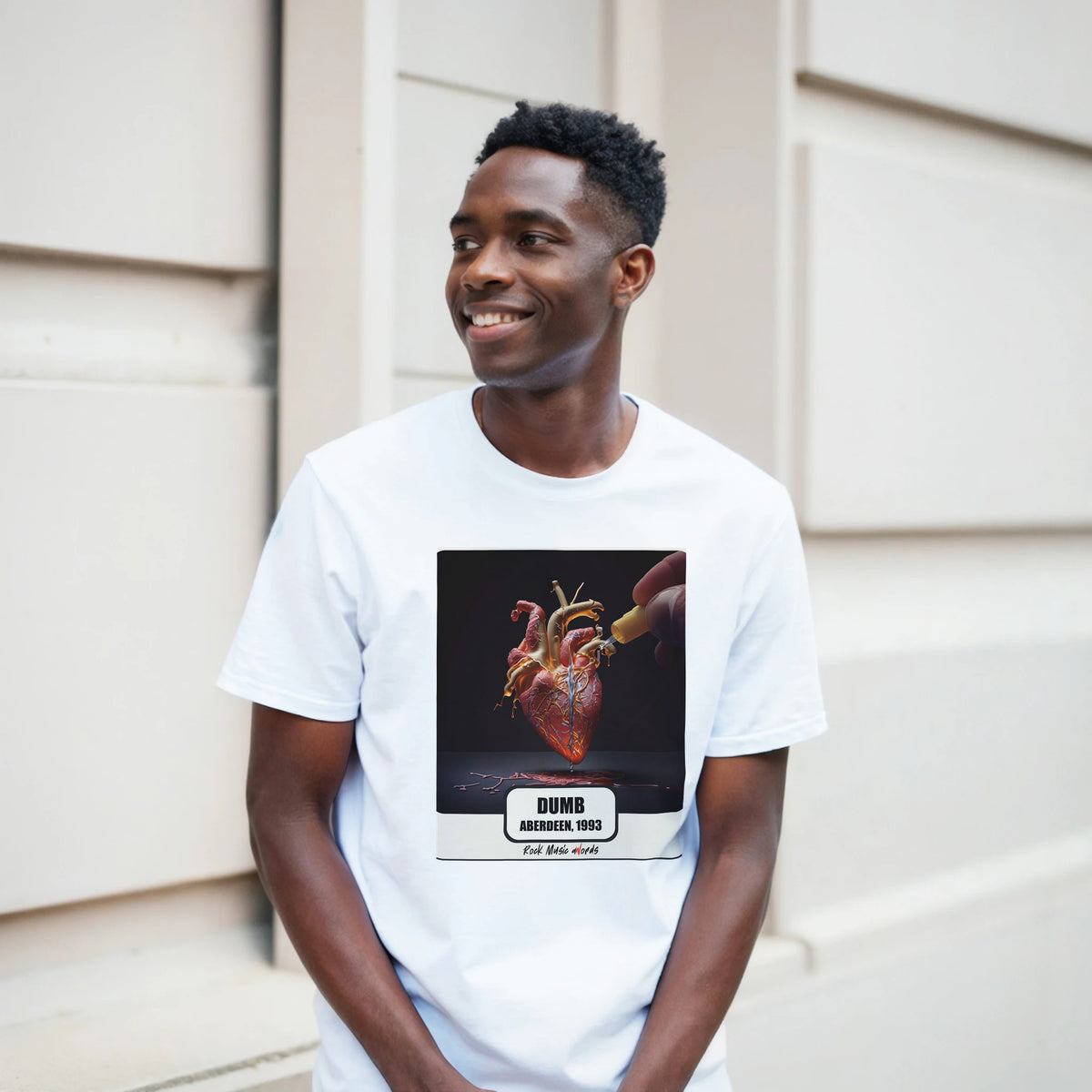 Ragazzo sorridente davanti a un muro chiaro a blocchi indossa la t-shirt bianca con la stampa Rock Art «Dumb – Aberdeen, 1993»