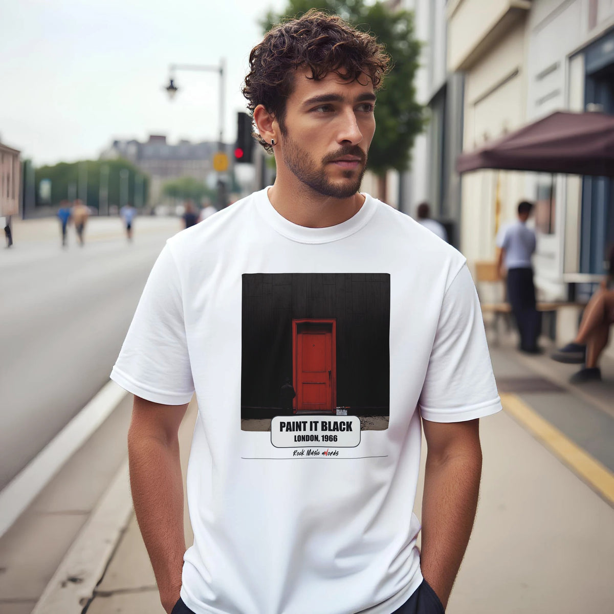 Uomo con capelli ricci e barba su un marciapiede di città indossa la t-shirt bianca con la stampa Rock Art «Paint It Black – London, 1966»