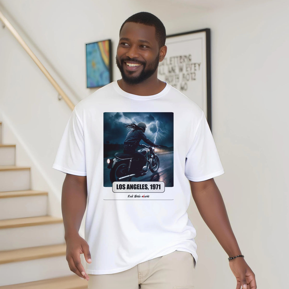 Uomo con la barba sorride vicino a una scala di legno, in casa, e indossa la t-shirt bianca con la stampa Rock Art «Riders on the Storm – Los Angeles, 1971»