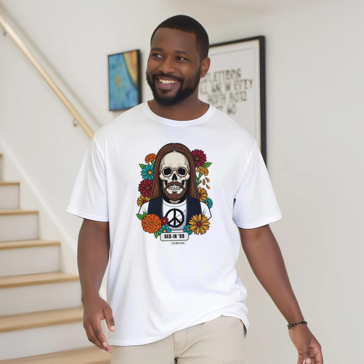Uomo con la barba sorride vicino a una scala di legno, in casa, e indossa la t-shirt bianca con la calavera Bed-In '69