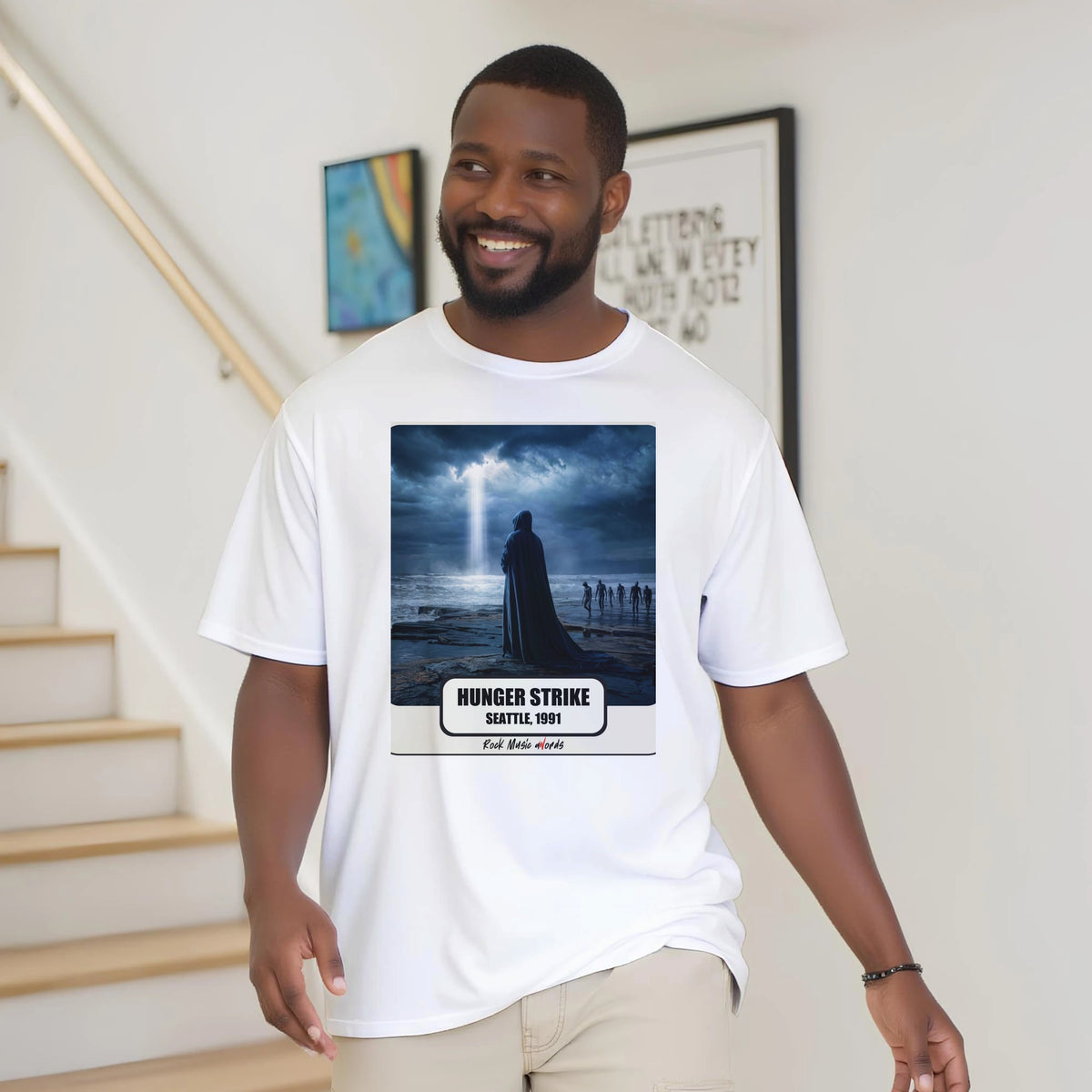 Uomo con la barba sorride vicino a una scala di legno, in casa, e indossa la t-shirt bianca con la stampa Rock Art «Hunger Strike – Seattle, 1991»