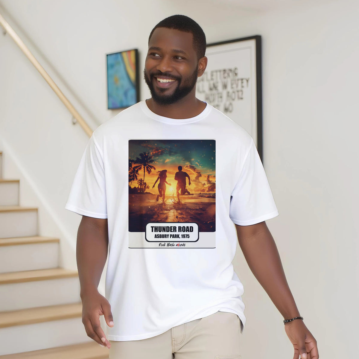 Uomo con la barba sorride vicino a una scala di legno, in casa, e indossa la t-shirt bianca con la stampa Rock Art «Thunder Road – Asbury Park, 1975»