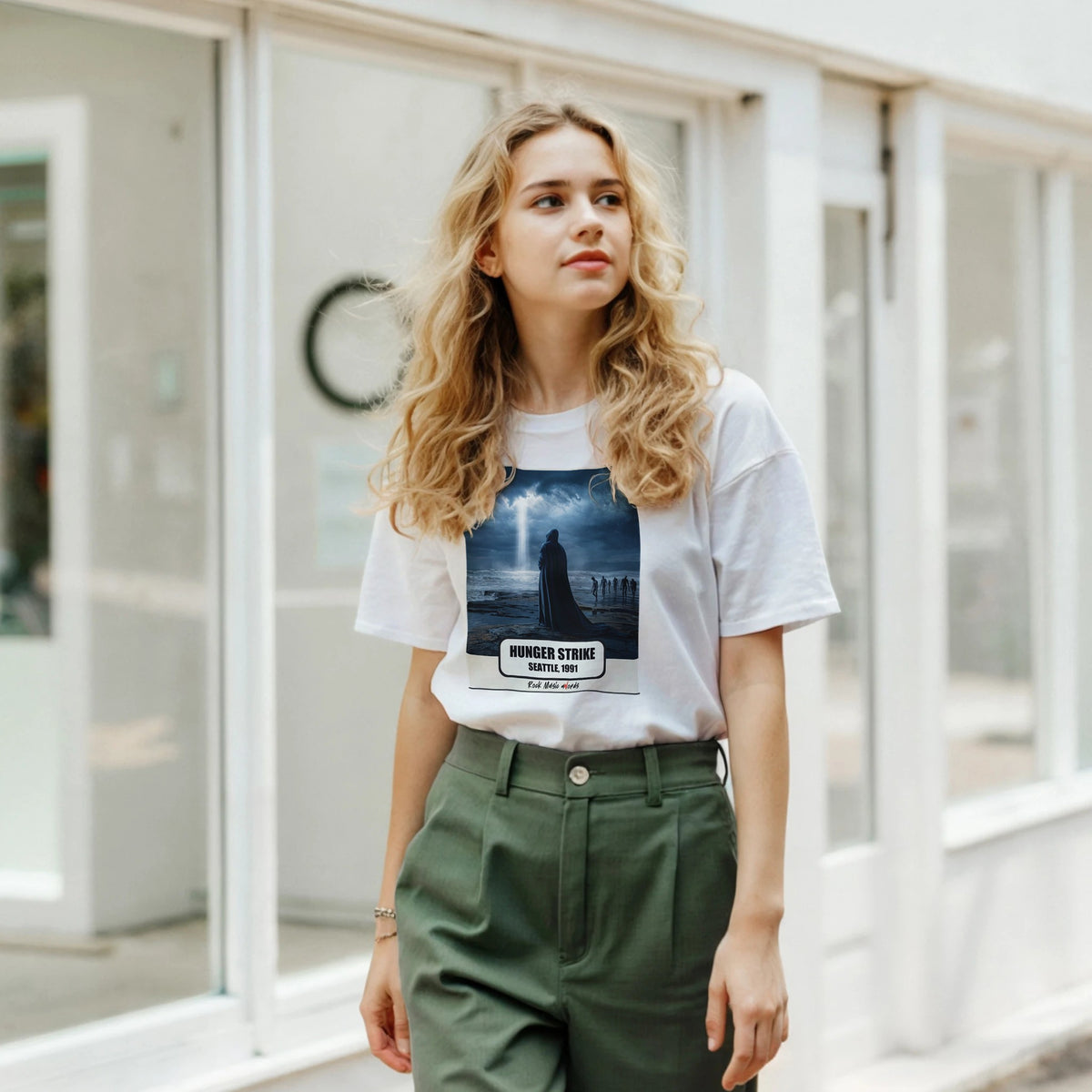 Ragazza bionda davanti a una facciata a vetrate indossa la t-shirt bianca con la stampa Rock Art «Hunger Strike – Seattle, 1991» e pantaloni verdi