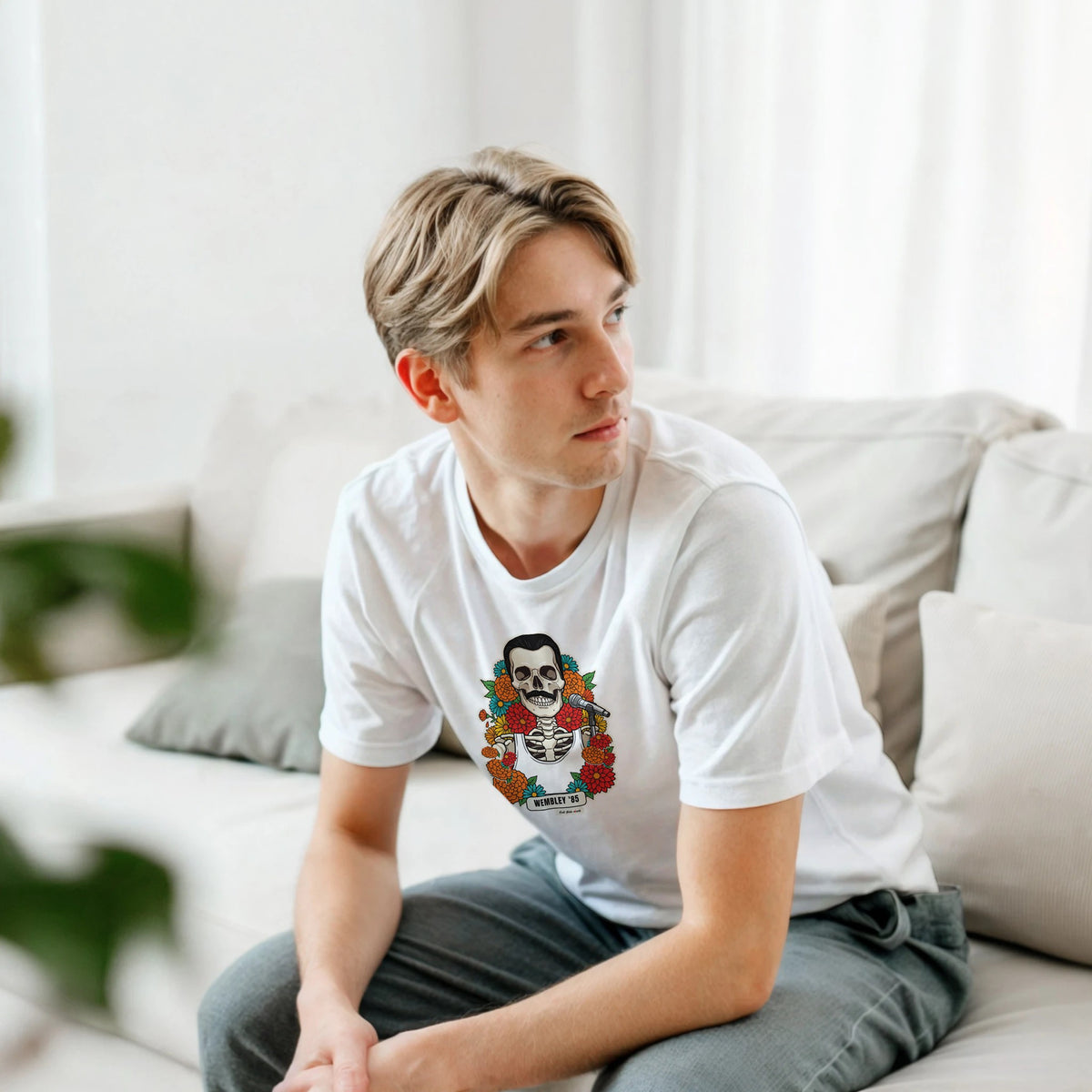 Ragazzo biondo seduto su un divano bianco, piegato in avanti, indossa la t-shirt bianca con la calavera Wembley '85