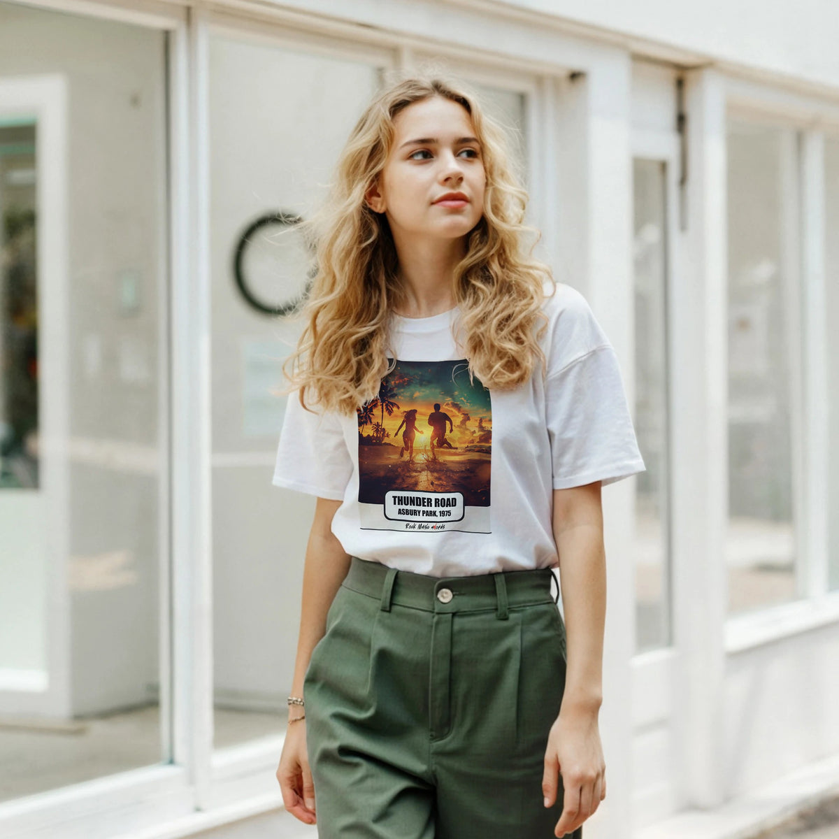 Ragazza bionda davanti a una facciata a vetrate indossa la t-shirt bianca con la stampa Rock Art «Thunder Road – Asbury Park, 1975» e pantaloni verdi
