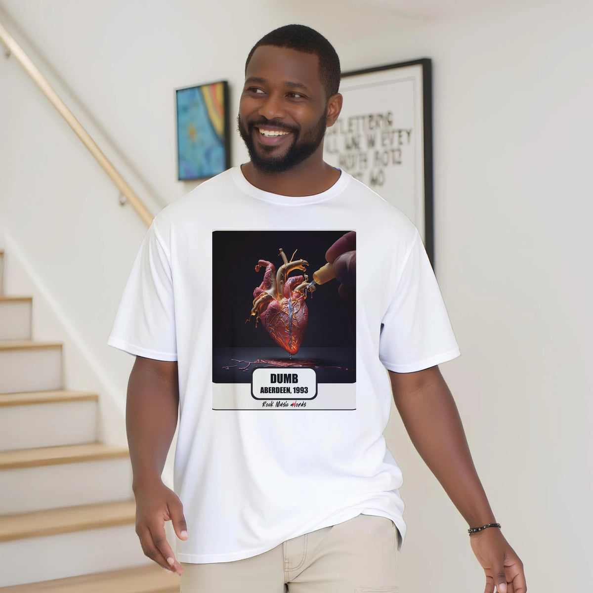 Uomo con la barba sorride vicino a una scala di legno, in casa, e indossa la t-shirt bianca con la stampa Rock Art «Dumb – Aberdeen, 1993»