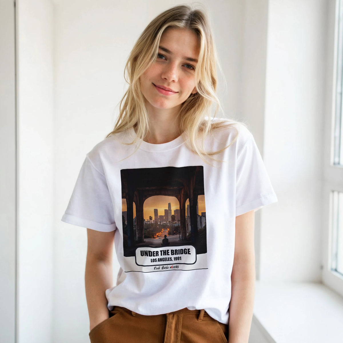 Ragazza bionda con le mani in tasca, accanto a una finestra, indossa la t-shirt bianca con la stampa Rock Art «Under the Bridge – Los Angeles, 1991»