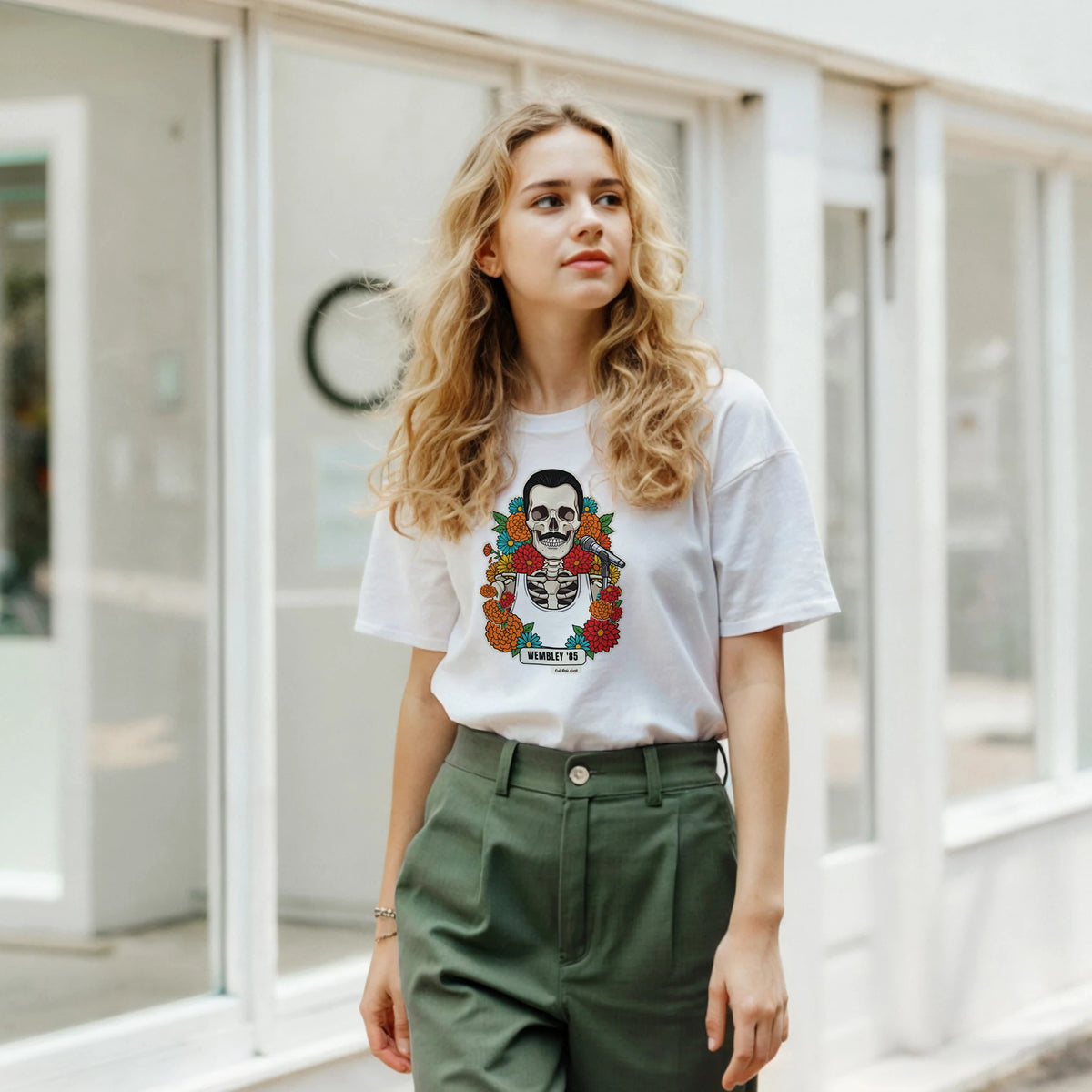 Ragazza bionda davanti a una facciata a vetrate indossa la t-shirt bianca con la calavera Wembley '85 e pantaloni verdi
