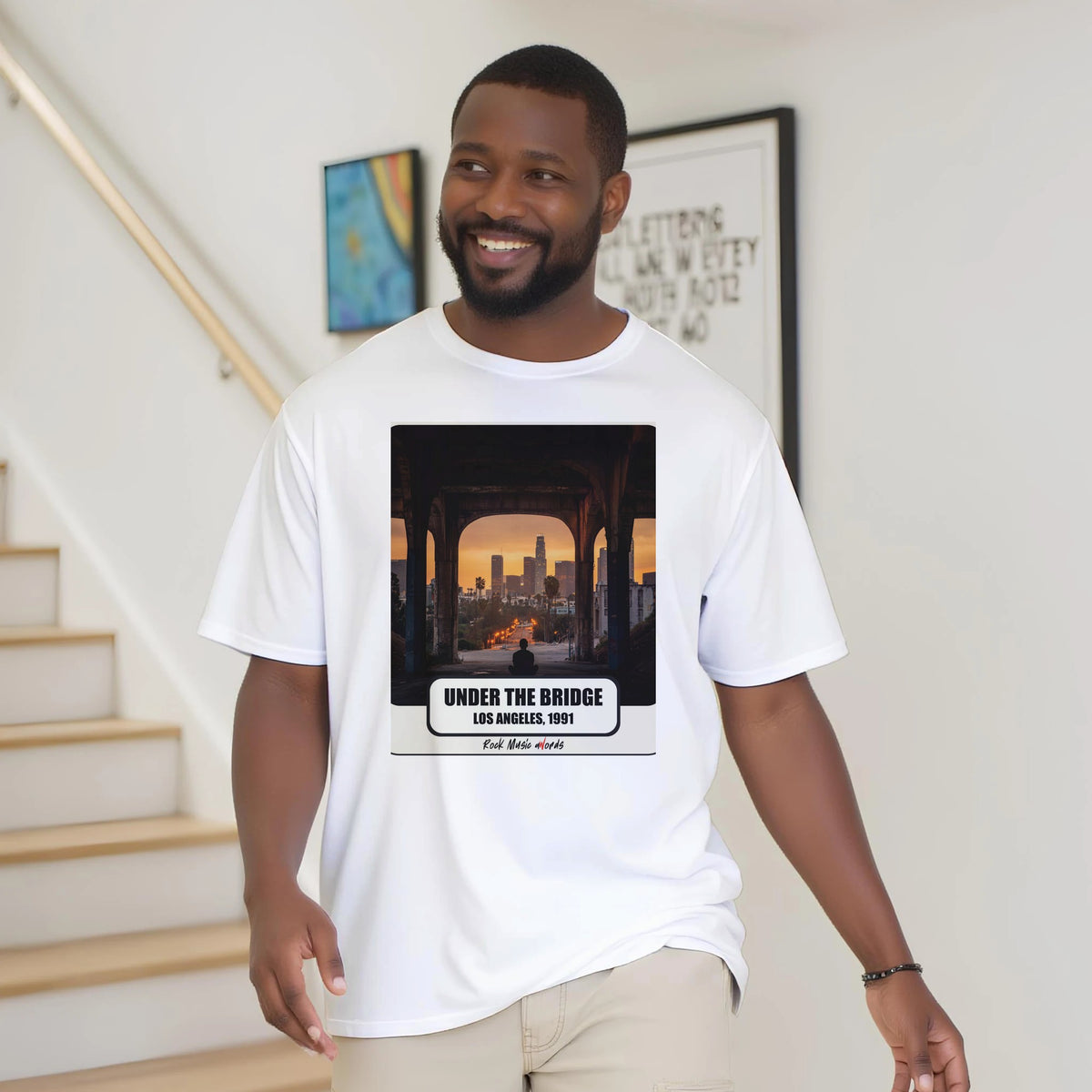 Uomo con la barba sorride vicino a una scala di legno, in casa, e indossa la t-shirt bianca con la stampa Rock Art «Under the Bridge – Los Angeles, 1991»