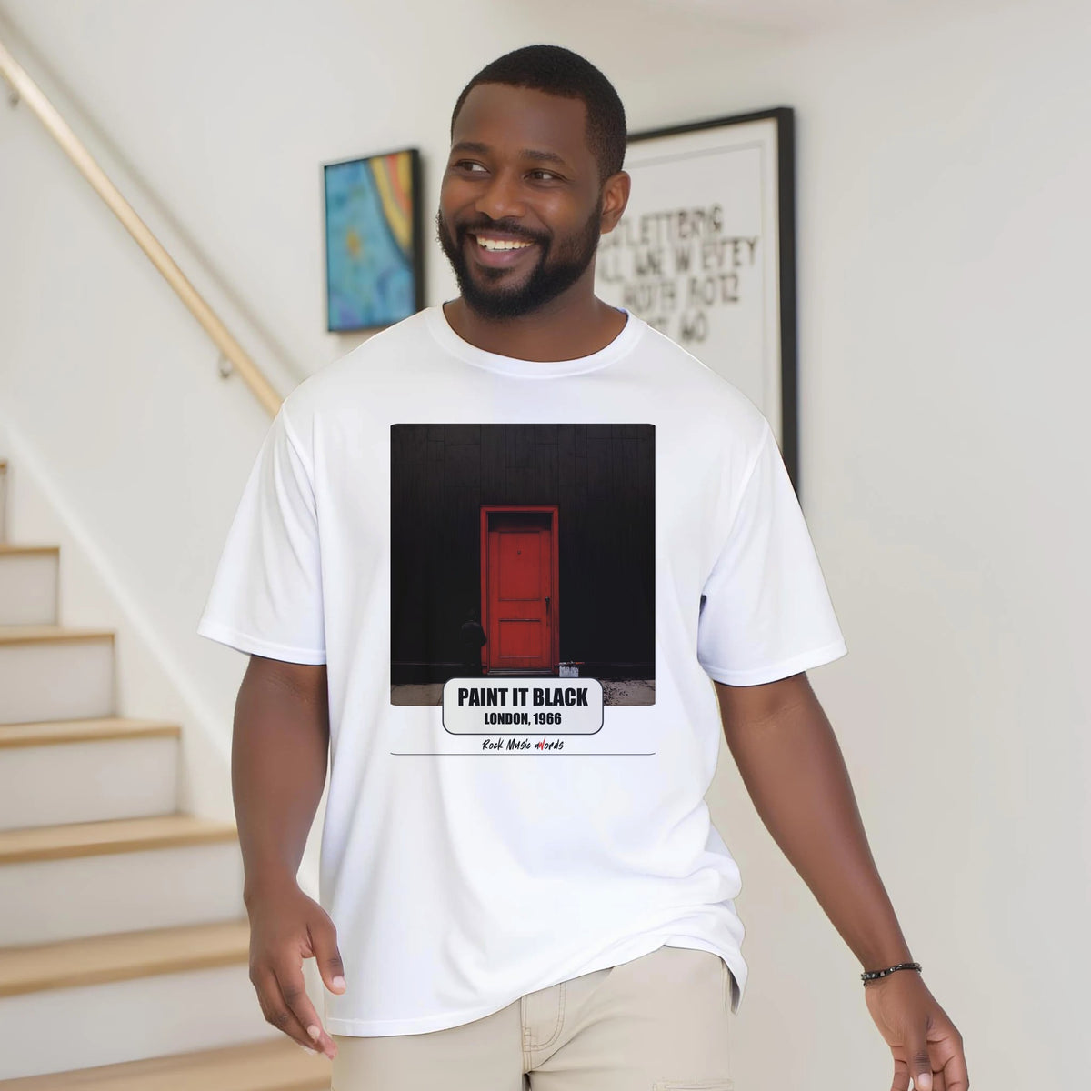 Uomo con la barba sorride vicino a una scala di legno, in casa, e indossa la t-shirt bianca con la stampa Rock Art «Paint It Black – London, 1966»