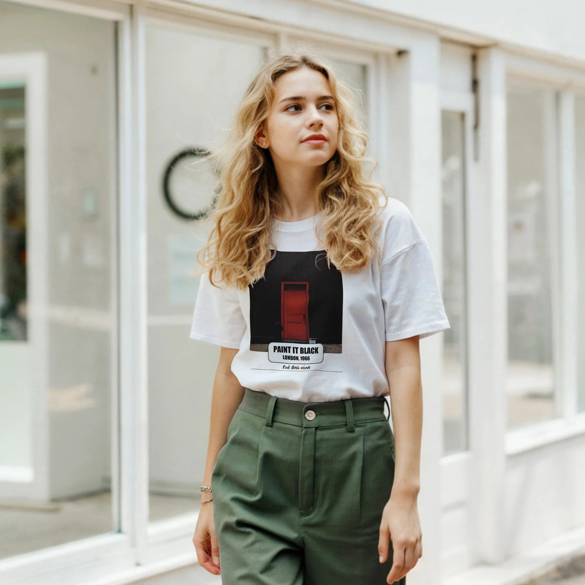 Ragazza bionda davanti a una facciata a vetrate indossa la t-shirt bianca con la stampa Rock Art «Paint It Black – London, 1966» e pantaloni verdi