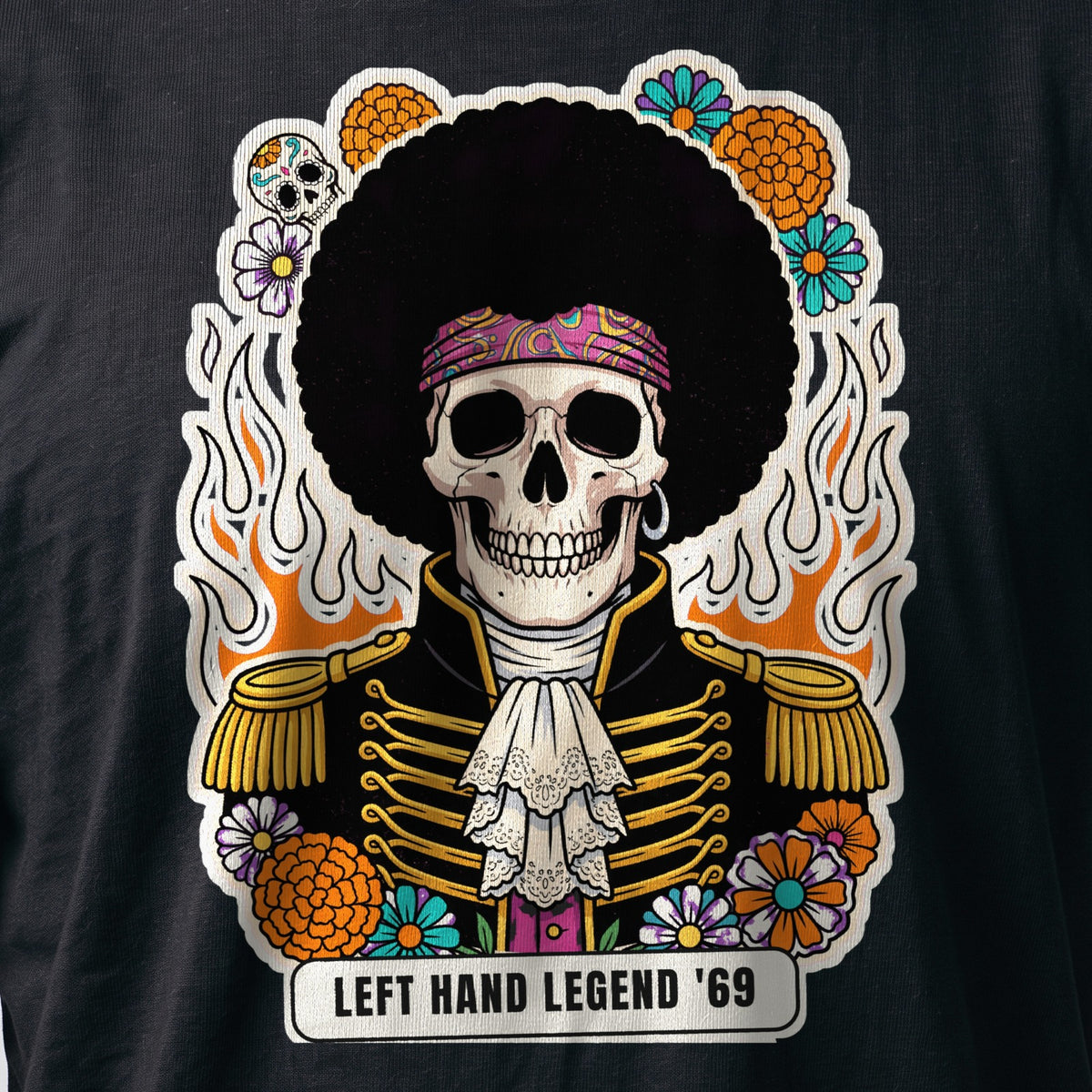 La stampa da vicino sulla t-shirt nera: la calavera Left Hand Legend con i capelli afro, la fascia, la giacca da ussaro e le fiamme, sul cotone (ricostruzione al computer)